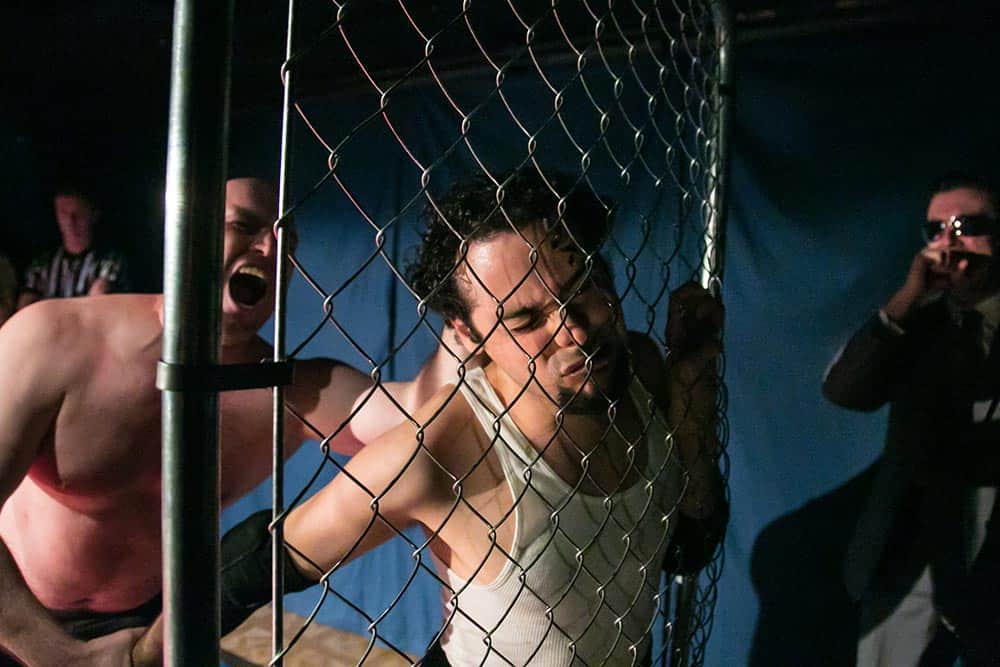 Production image from "As You Like It" at Impact Theatre: a cage fight with someone getting his face smashed against a fence
